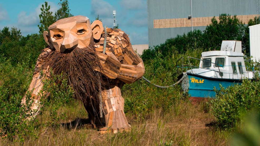 A large wooden troll sculpture in a grassy area, with a boat tied nearby, surrounded by trees and buildings.